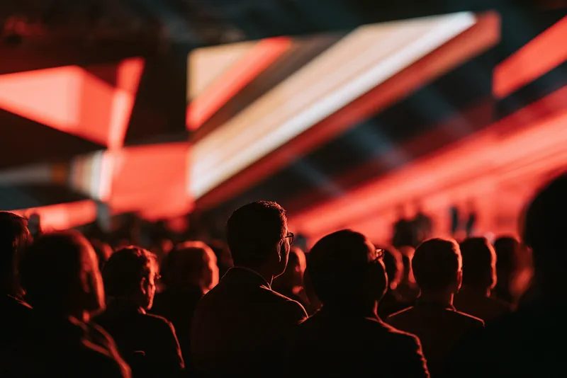 Opening Titles — a conference audience in silhouette beneath a vast angled screen of red, coral and white geometric shapes