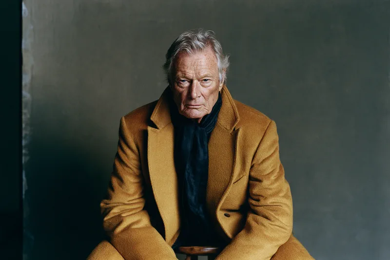 Sunday Best — portrait of a silver-haired man in an ochre wool overcoat and dark scarf, seated against a painted grey backdrop in window light